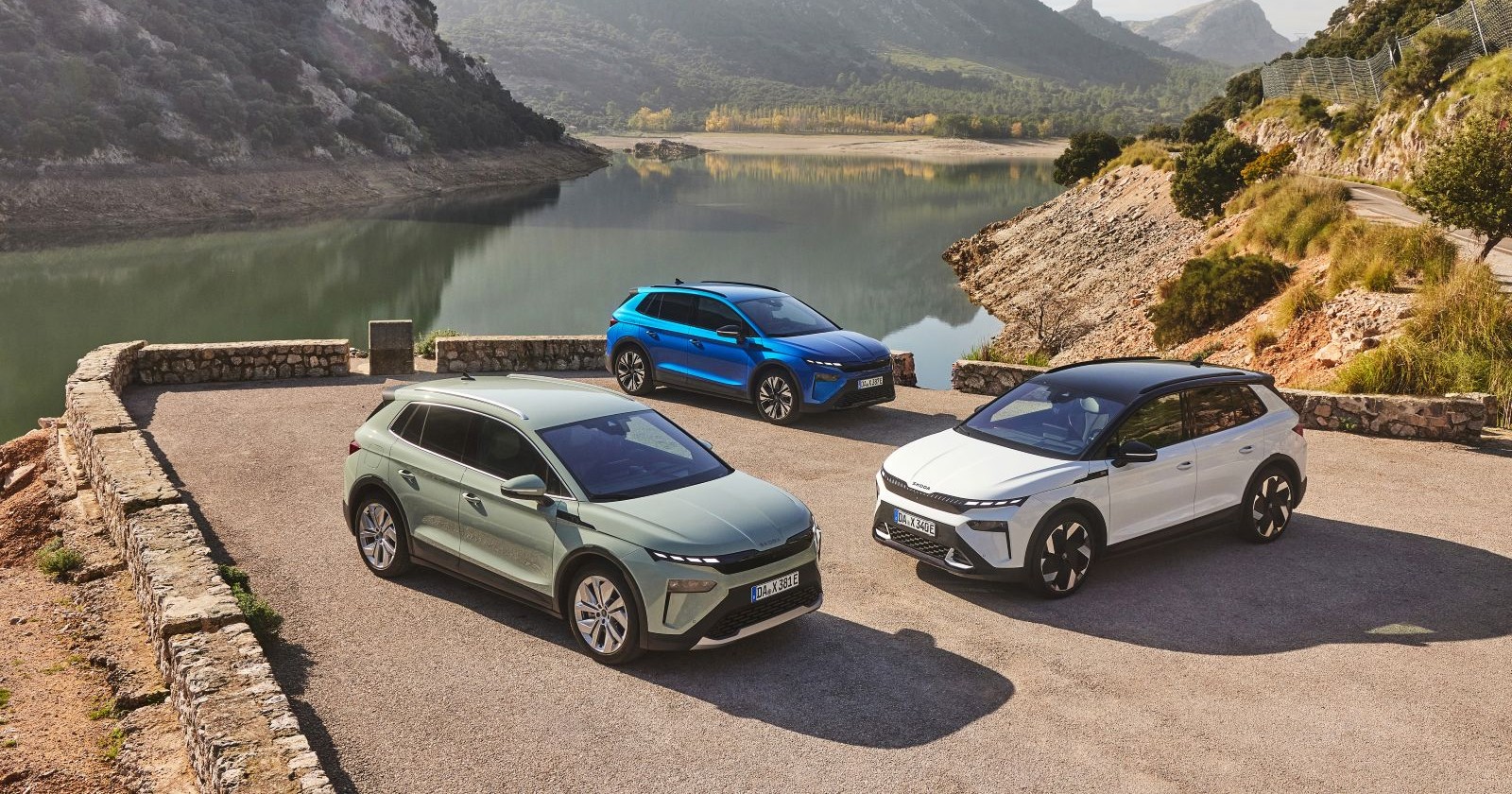 Drei Skoda Elroq, in den Farben Weiß, Blau und Grün, parken am Rande eines Stausees. Die karge Landschaft erinnert an Südeuropa.