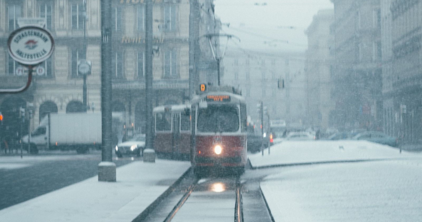 Eine rote Wiener Straßenbahn fährt bei starkem Schneefall durch eine verschneite Innenstadtstraße, die Gleise und Fahrbahnen sind mit Schnee bedeckt./ Starker Schneefall legt den Straßenbahnverkehr in Wien teilweise lahm