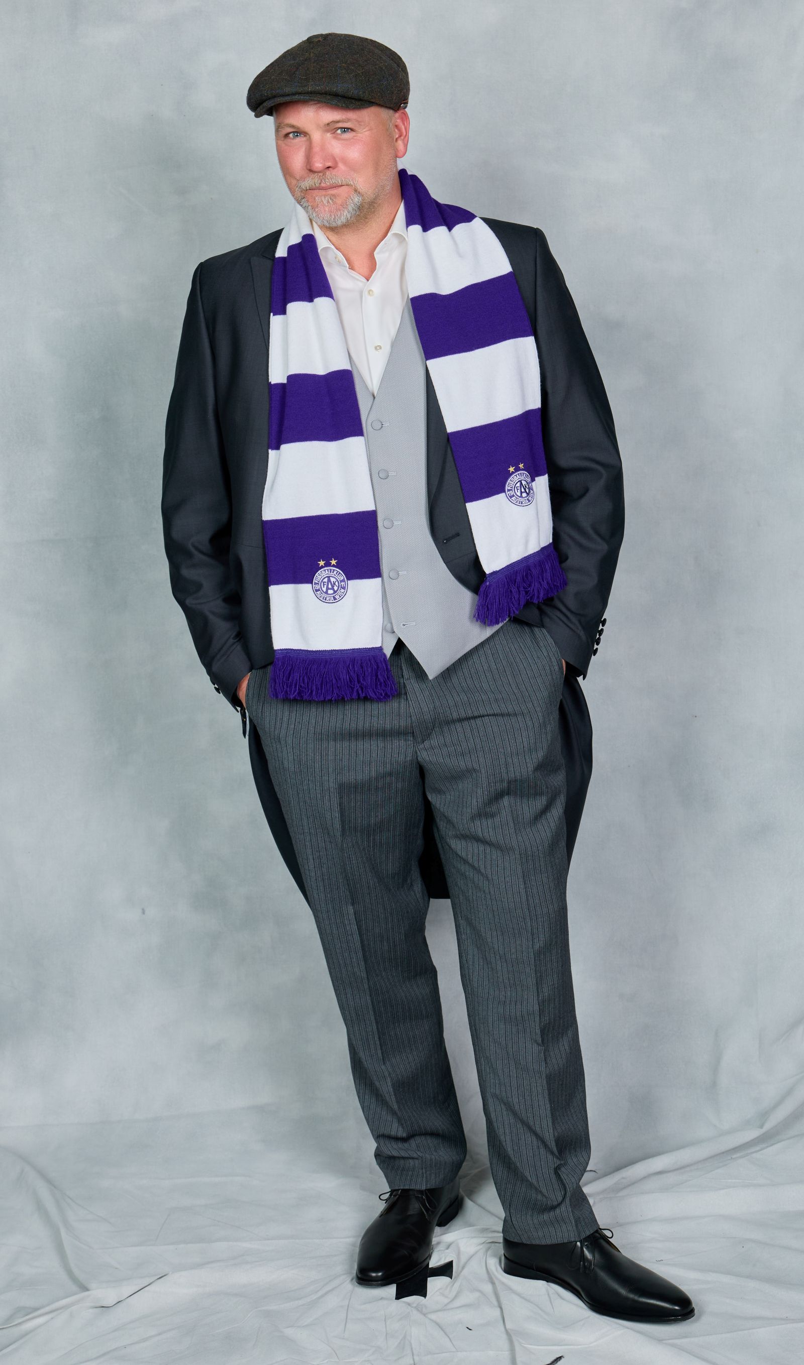 Thomas Stipsits steht im Anzug mit violett-weißem Schal vor neutralem Hintergrund. Er spielt im ORF-Dokudrama „Benko – Size does matter“ den Unternehmer René Benko.