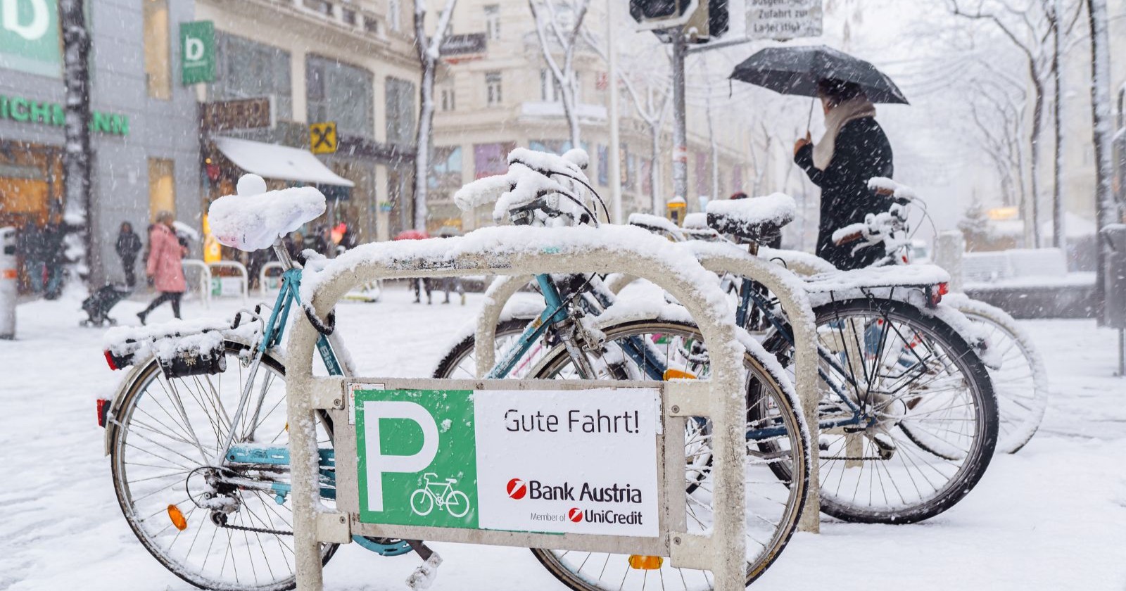 Verschneite Fahrräder an einem Abstellplatz in einer Wiener Einkaufsstraße, während dichter Schneefall und Passanten mit Schirmen das winterliche Stadtbild prägen.