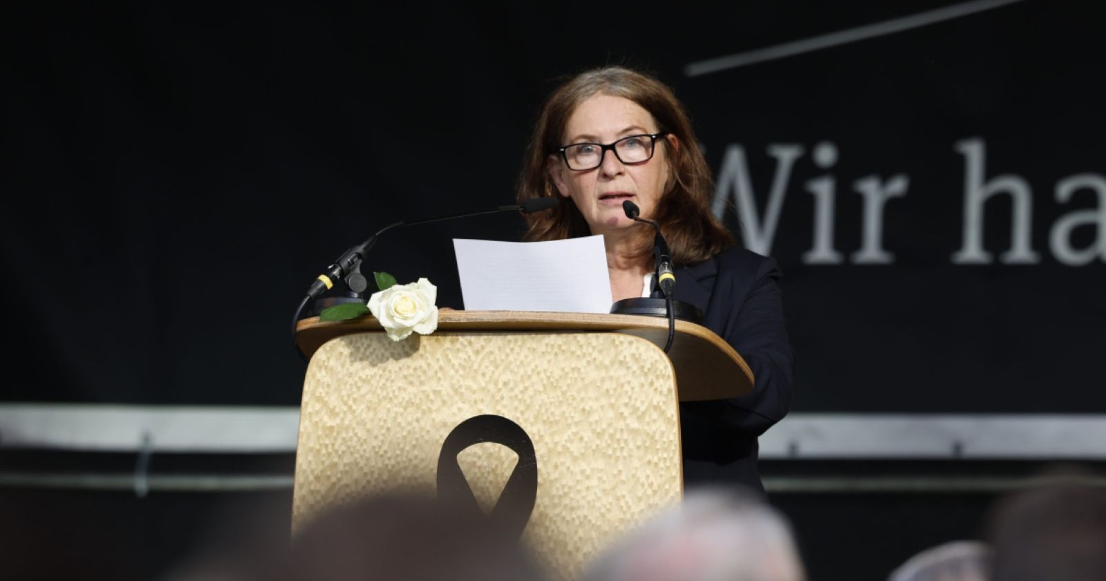 Gedenkveranstaltung nach Amoklauf in Graz: Frau spricht am Rednerpult mit schwarzer Trauerschleife und weißer Rose/Ein sTipendium soll an Lehrerin Dorit Wiener erinnern