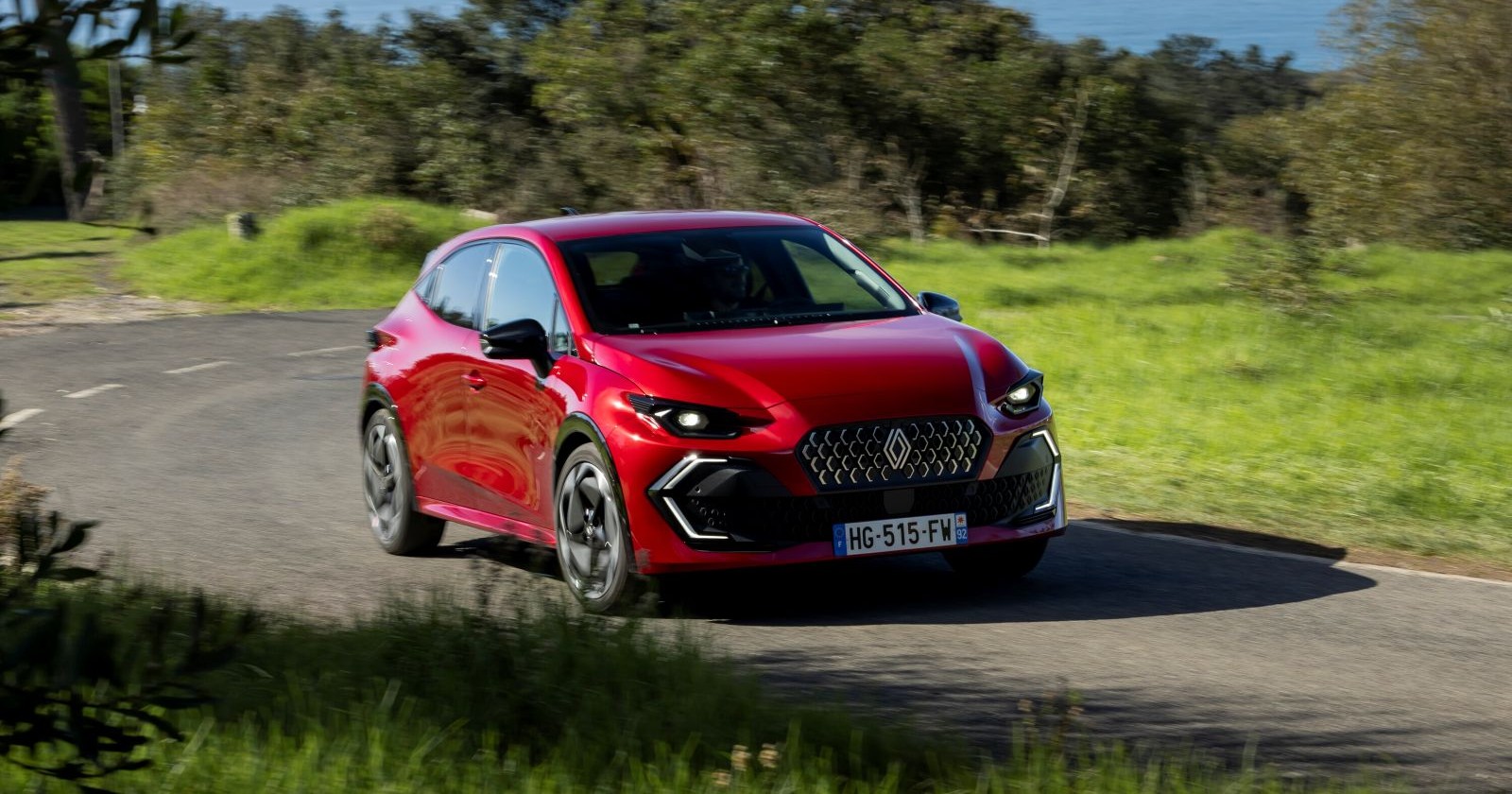 Ein roter Renault Clio fährt auf einer Landstraße. Neben der Straße ist eine grüne Wiese zu sehen, im Hintergrund ein Laubwald.