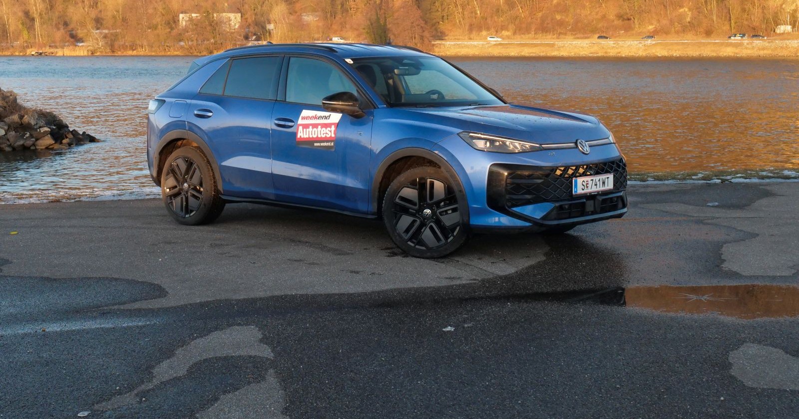 Ein hellblauer VW T-Rock parkt am Rande eines Gewässern. Am anderen Ufer sind Bäume ohne Laub zu sehen, wahrscheinlich ist es Winter.