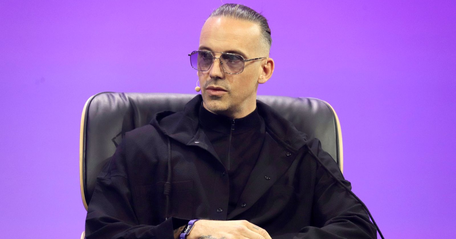 RAF Camora sitzt auf einer Bühne bei einem Interview oder Panel, trägt schwarze Kleidung und eine Sonnenbrille und spricht über seine Rückkehr von Dubai nach Wien nach den Iran-Angriffen.