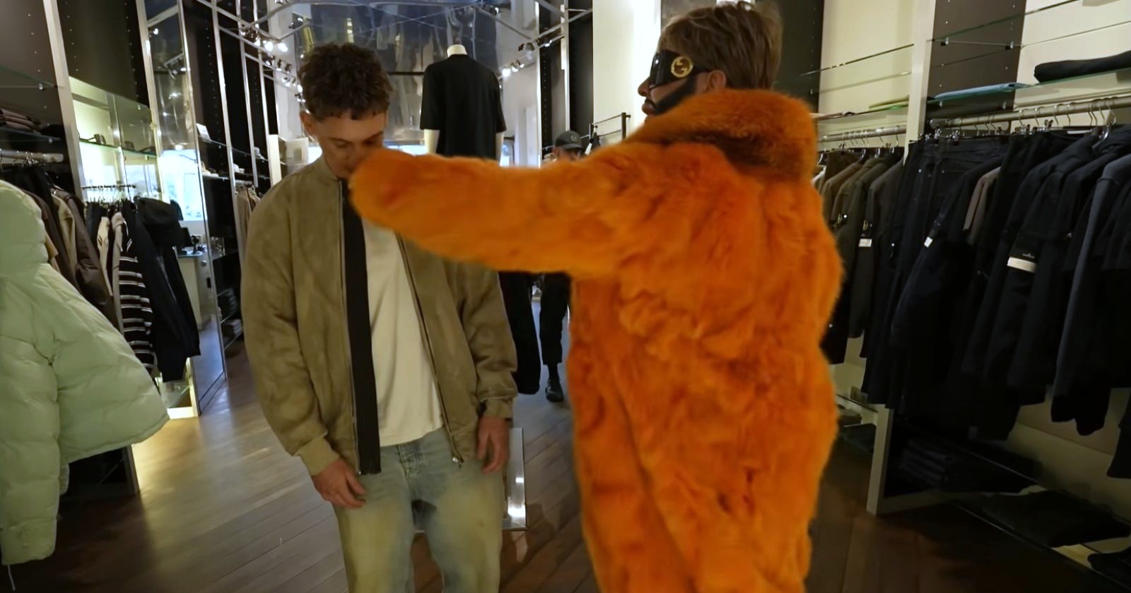 Harald Glööckler in einem auffälligen orangefarbenen Pelzmantel holt in einem Modegeschäft zum Schlag gegen einen jungen Mann aus, während Kleidung an den Wänden hängt.