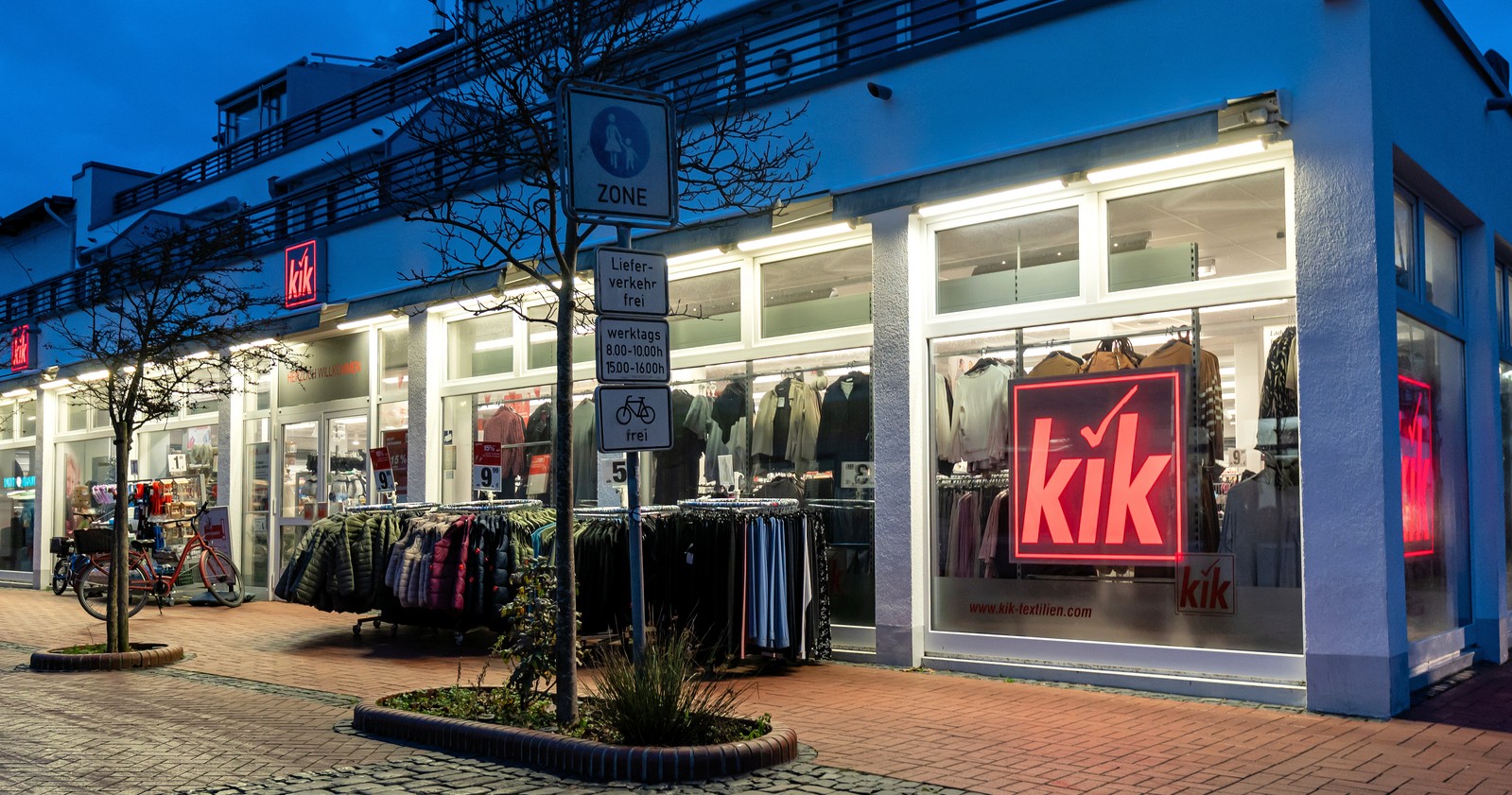 Beleuchtete KiK-Filiale in einer Einkaufsstraße mit Kleidung vor dem Geschäft und sichtbarem KiK-Logo im Schaufenster; Symbolbild für die Schließungen von KiK-Filialen