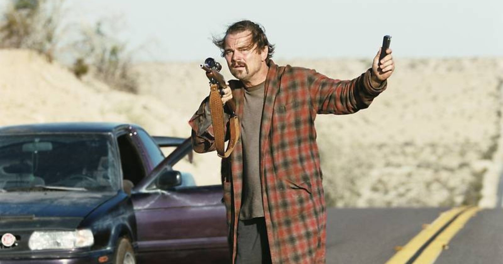 Leonardo DiCaprio in One Battle After Another mit Gewehr neben Auto auf Wüstenstraße – Szene aus dem Oscar-Film, der online gestreamt werden kann.