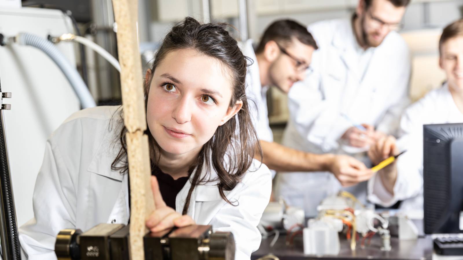 Junge Frau arbeitet an einem Gerät im Labor, während im Hintergrund weitere Personen gemeinsam experimentieren