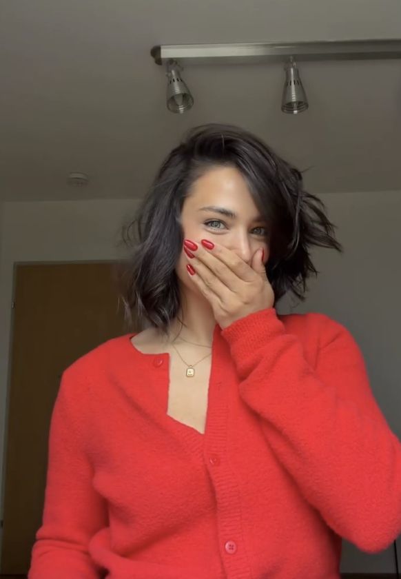 Amira Aly mit kurzem Bob und rotem Pullover reagiert überrascht auf ihre neue Frisur