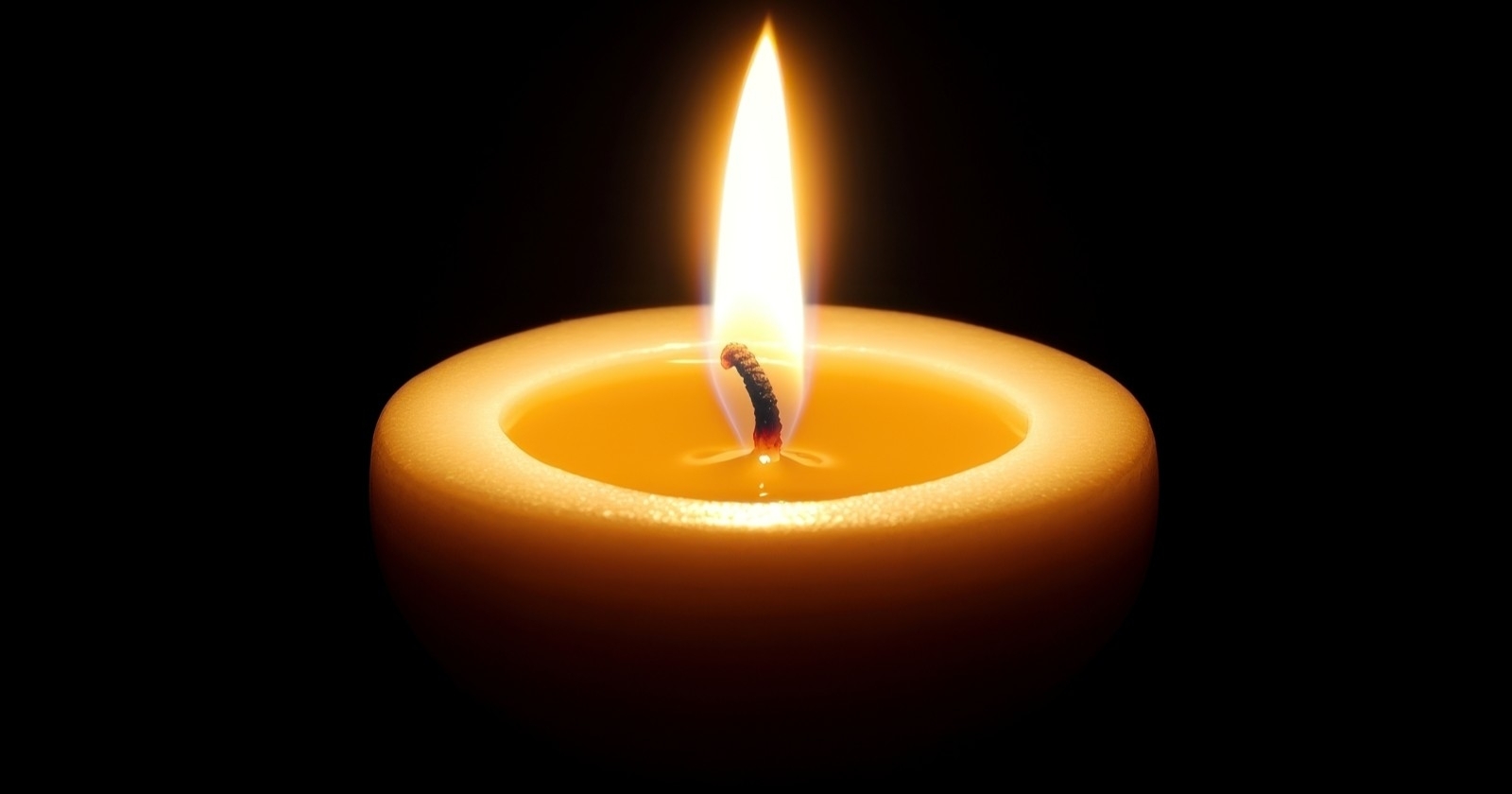 Eine einzelne, helle Kerze sitzt in der Mitte eines schwarzen Hintergrundes, deren warme gelbe Flamme einen sanften Schein auslöst.