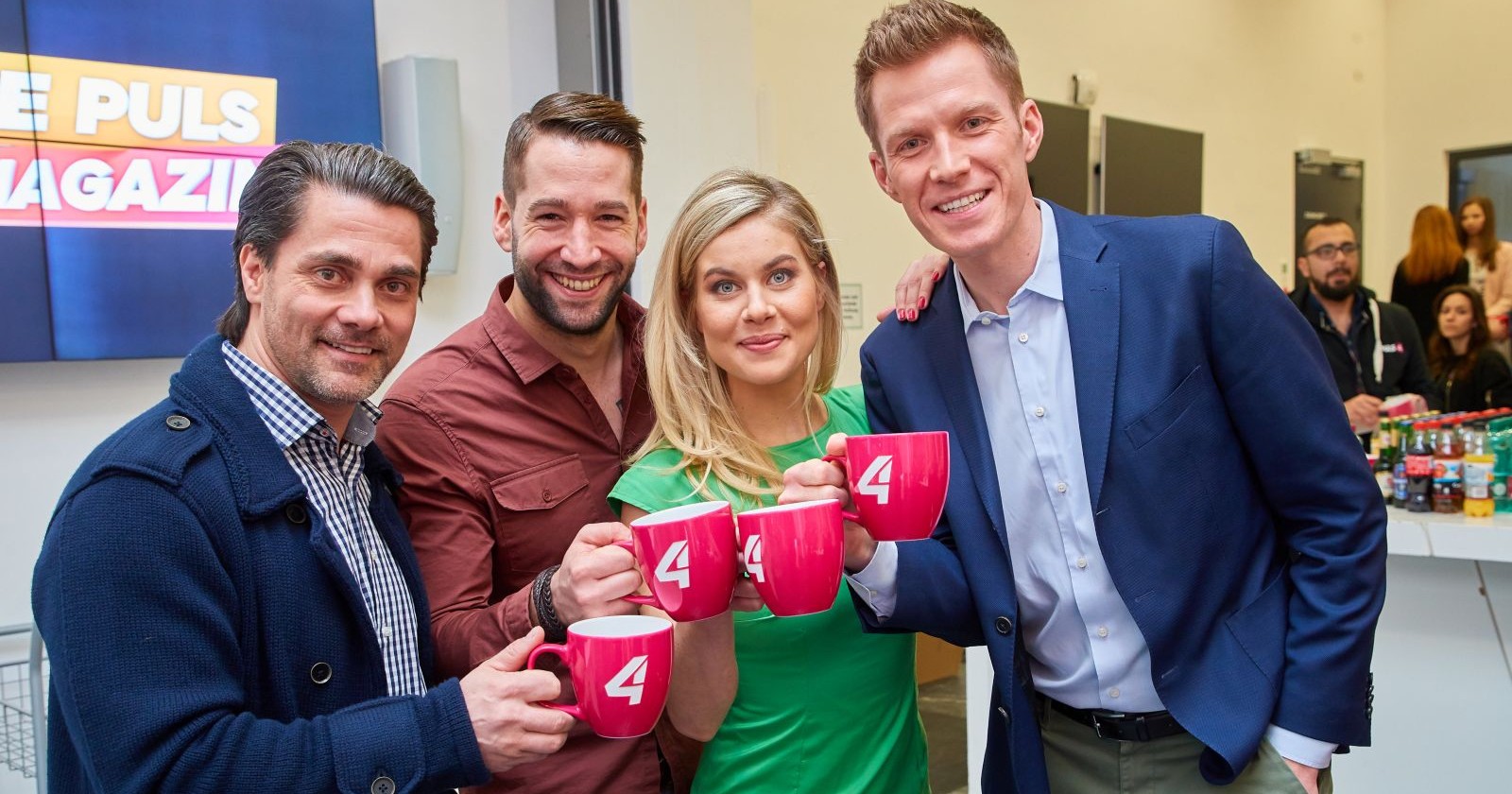 Dori Bauer schwanger: Puls4-Moderatorin mit Kollegen im Studio, hält Tasse mit „4“-Logo und lächelt in die Kamera