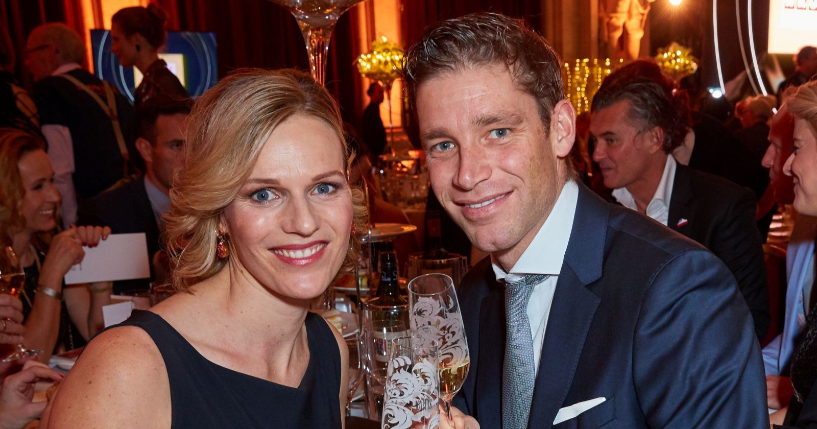 Marlies und Benni Raich bei einer festlichen Abendveranstaltung, elegant gekleidet mit Champagnergläsern in der Hand, lächelnd in die Kamera