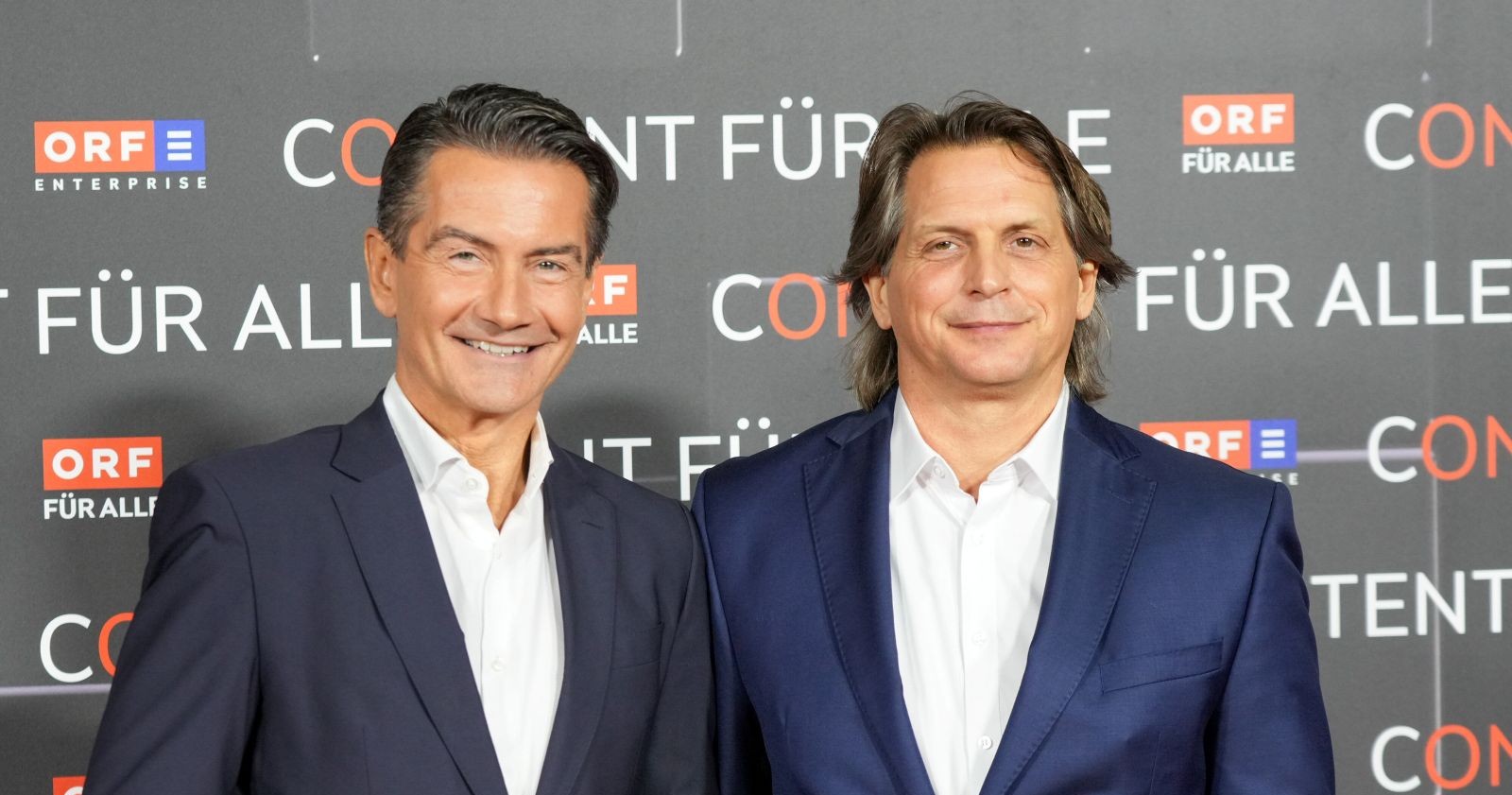 Oliver Böhm steht gemeinsam mit einem weiteren Manager vor einer ORF-Enterprise-Wand und blickt in die Kamera. Das Bild zeigt den ORF-Manager im Anzug im Kontext der aktuellen Vorwürfe und seiner Beurlaubung.