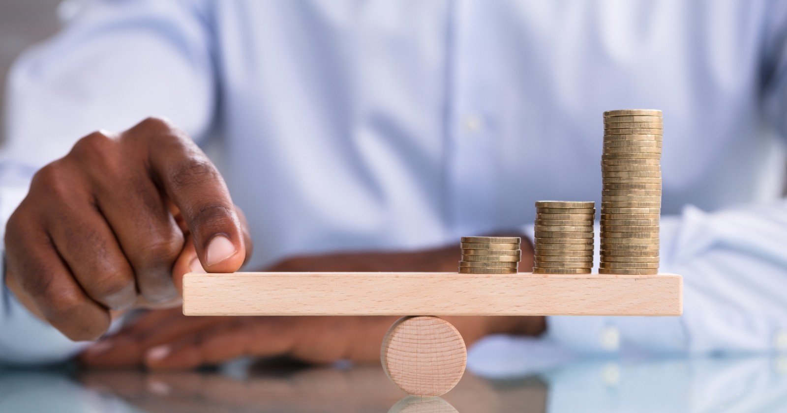 Nahaufnahme eines Geschäftsmannes, der den Münzstapel auf einer hölzernen Wippe über dem reflektierenden Schreibtisch balanciert.