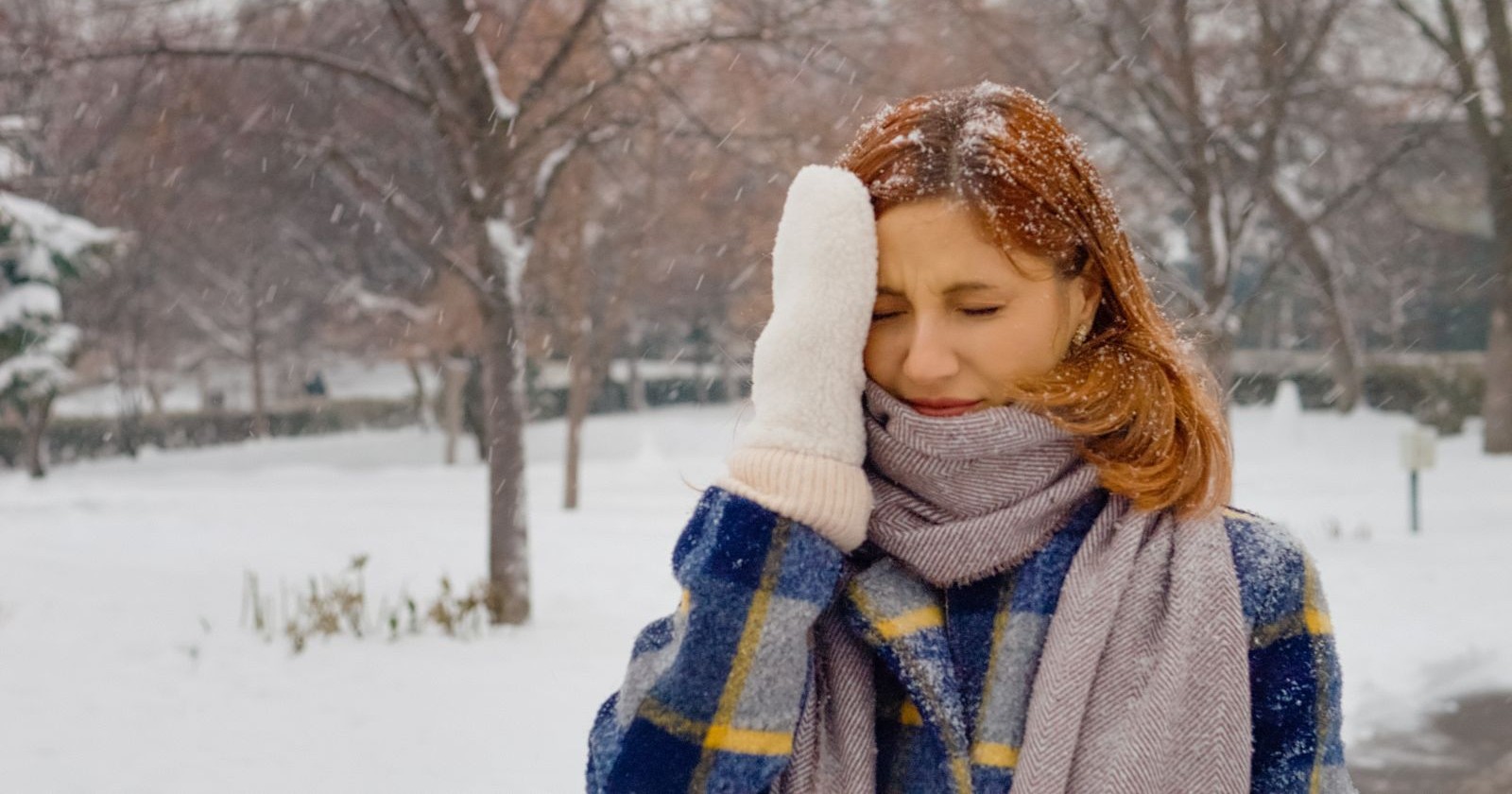 Frau greift sich in Winterkleidung an ihren Kopf, Schnee liegt im Hintergrund