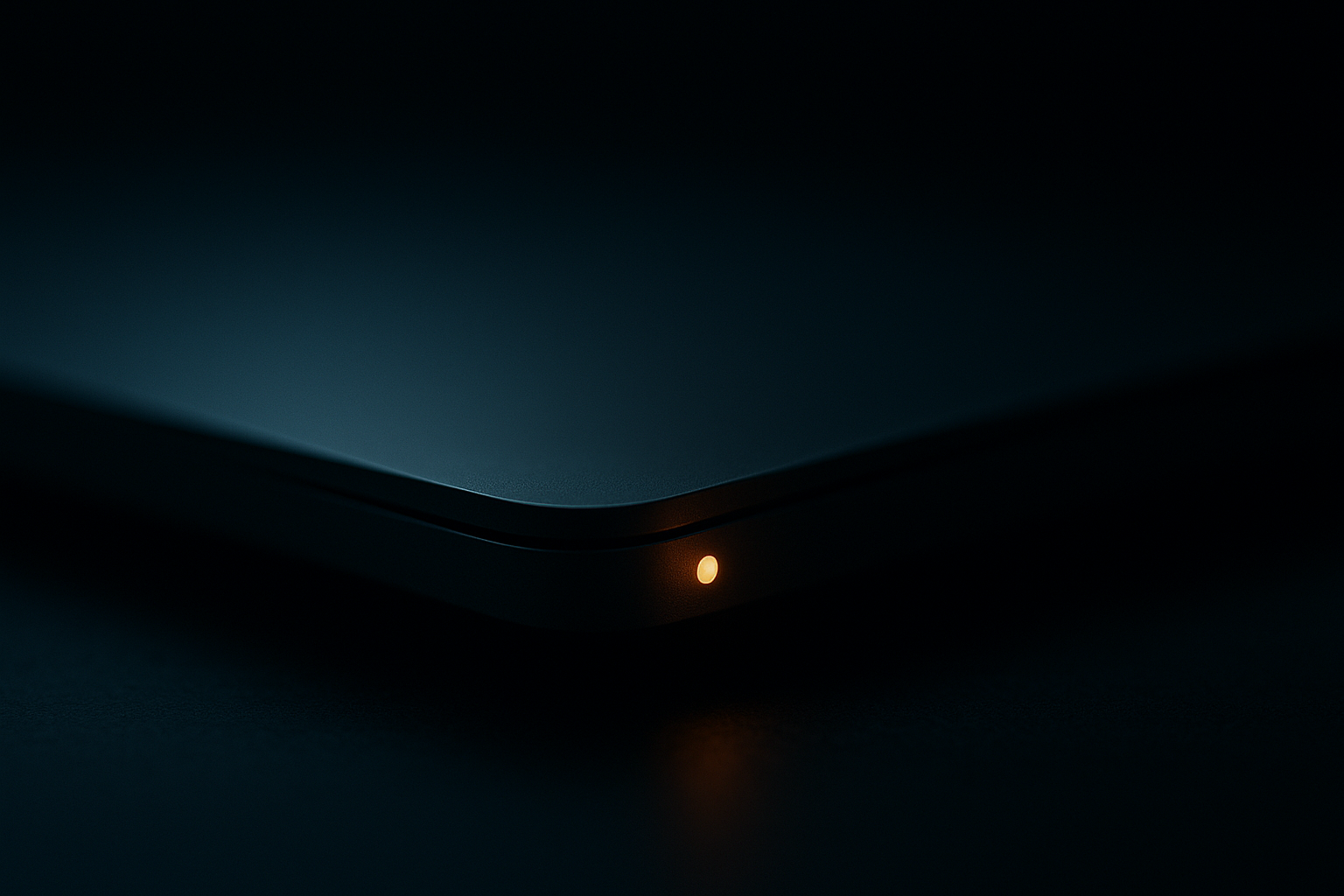 A moody close-up product shot of a laptop chassis edge in near-darkness, focused on a small illuminated amber warning indicator that glows confidently — even though everything around it is cool and calm. Cold slate-blue ambient light vs. the lone warm amber dot. Shallow depth of field, cinematic, minimal, editorial tech aesthetic. No text, no logos, no readable screens. 16:9.